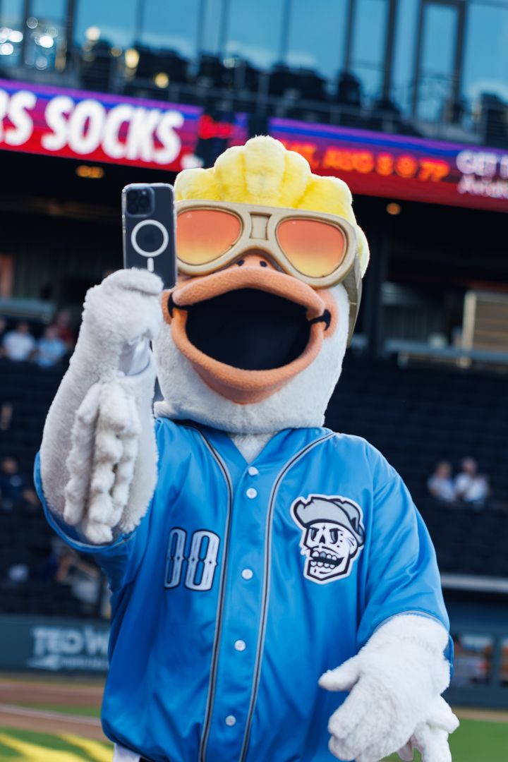 Baseball mascot dressed in blue jersey poses for selfie with phone at a minor league ballpark.