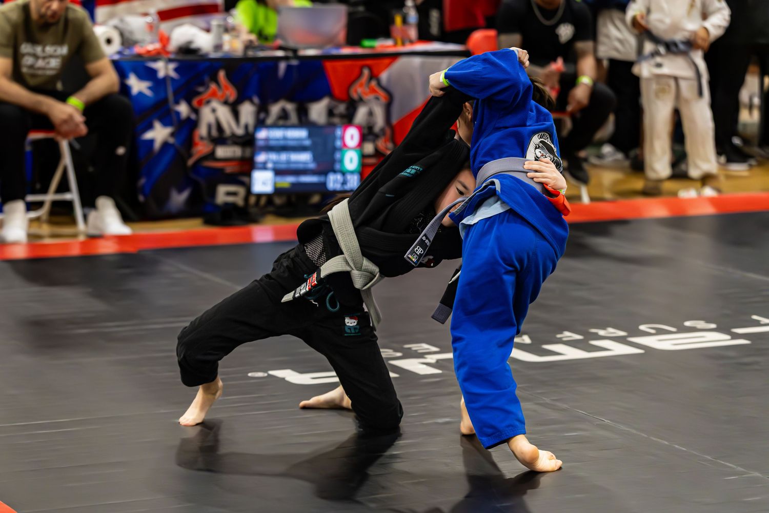 Two competitors in gi uniforms sparring during a Brazilian Jiu-Jitsu tournament on a black mat.