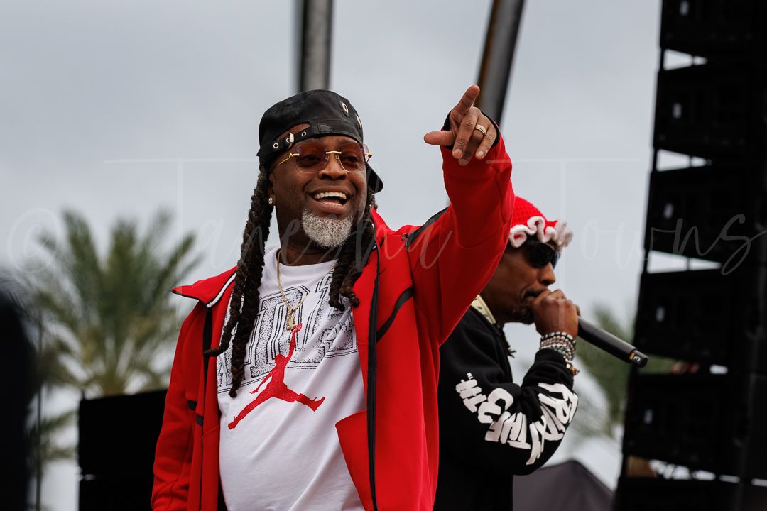 Performer in red Jordan jacket and dreads points finger on outdoor stage during live music event.