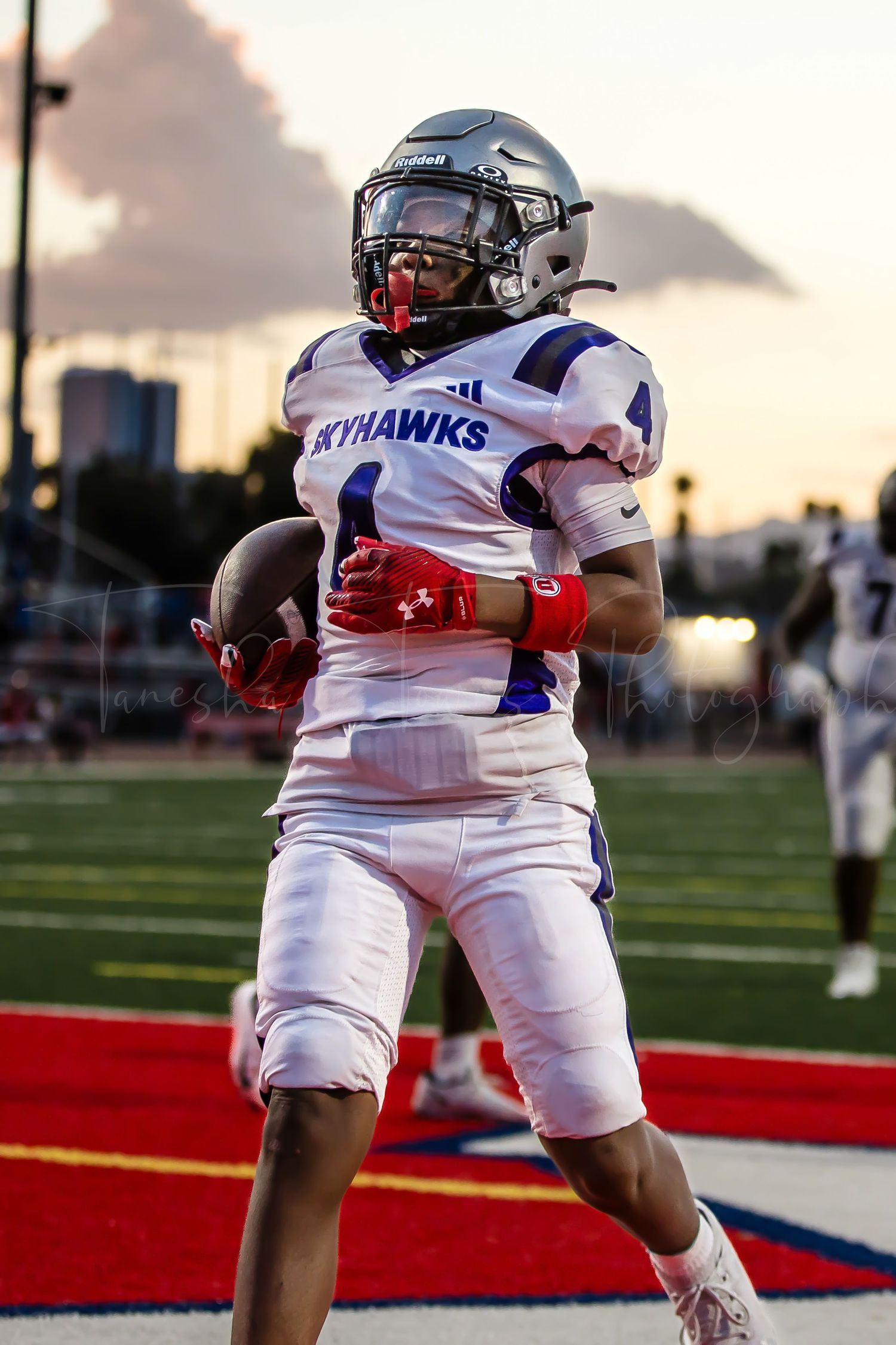 A Skyhawks football player in white uniform runs on the field at sunset.