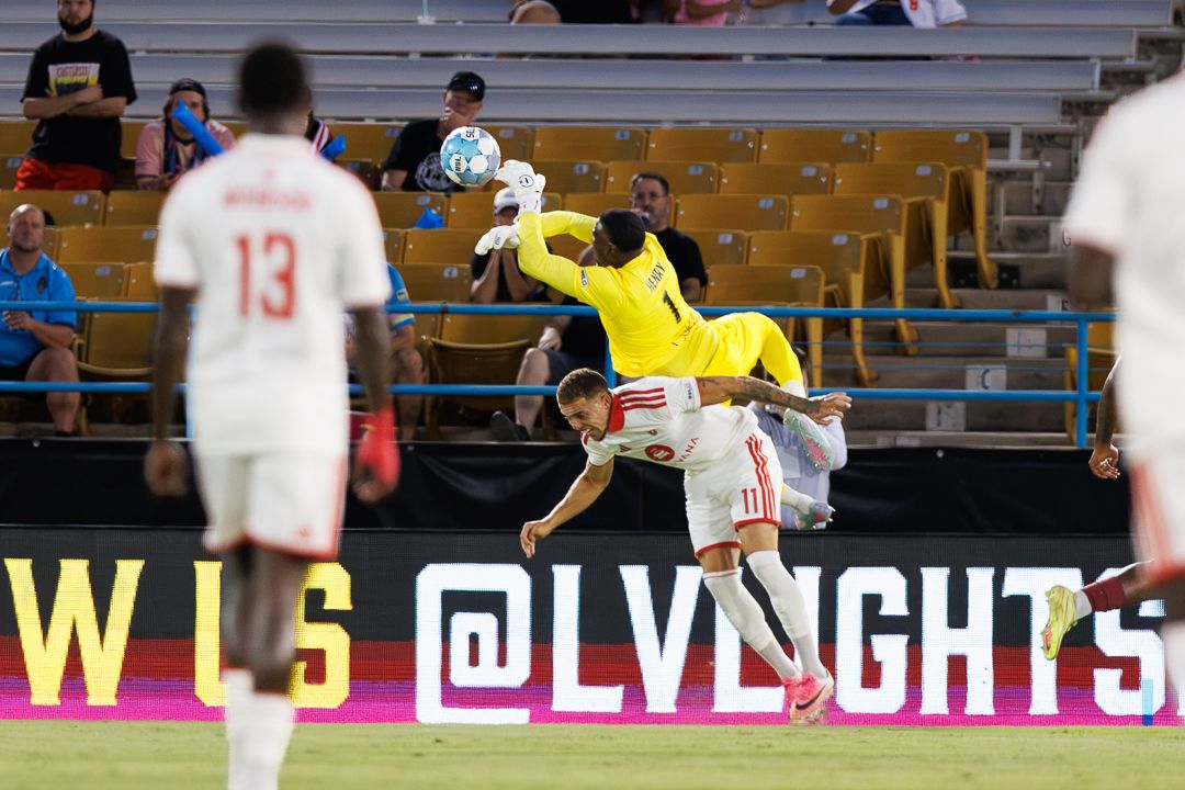 Goalkeeper leaps high to catch soccer ball during competitive match at LV Lights stadium under bright lights.