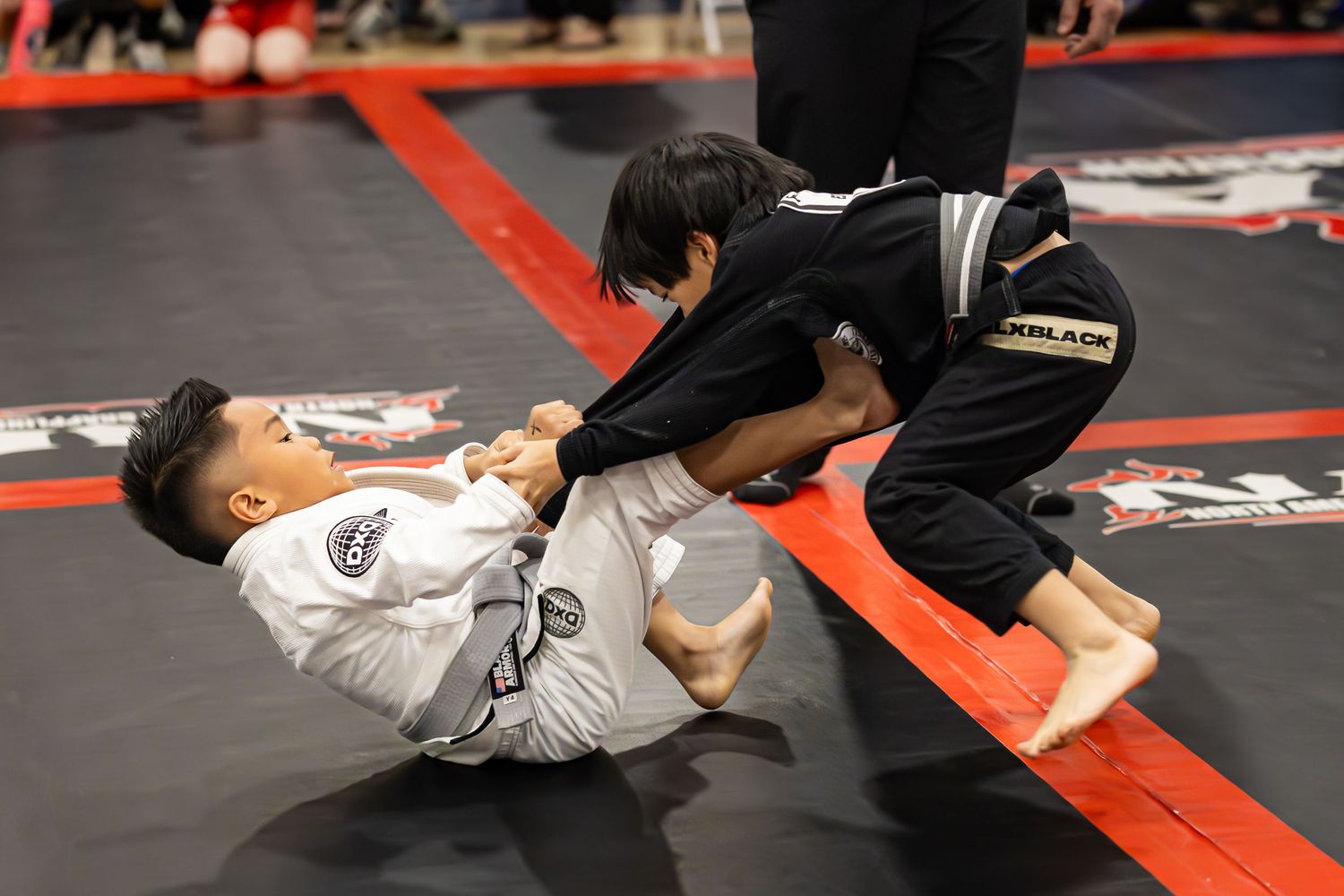 Two children competing in a Brazilian jiu-jitsu match on a black mat during a martial arts tournament.