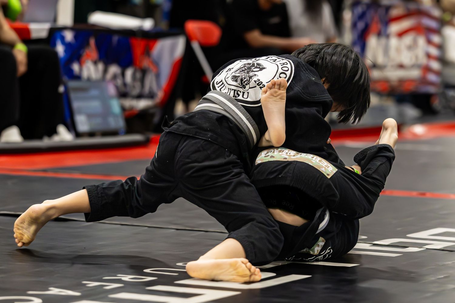 Two competitors in black gi uniforms grappling on a mat during a NASA jiu-jitsu tournament match.