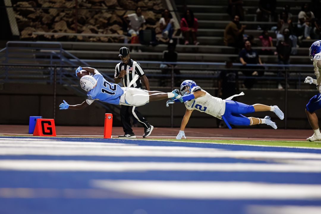 A football player in light blue dives into the end zone for a touchdown while a defender attempts to tackle him.