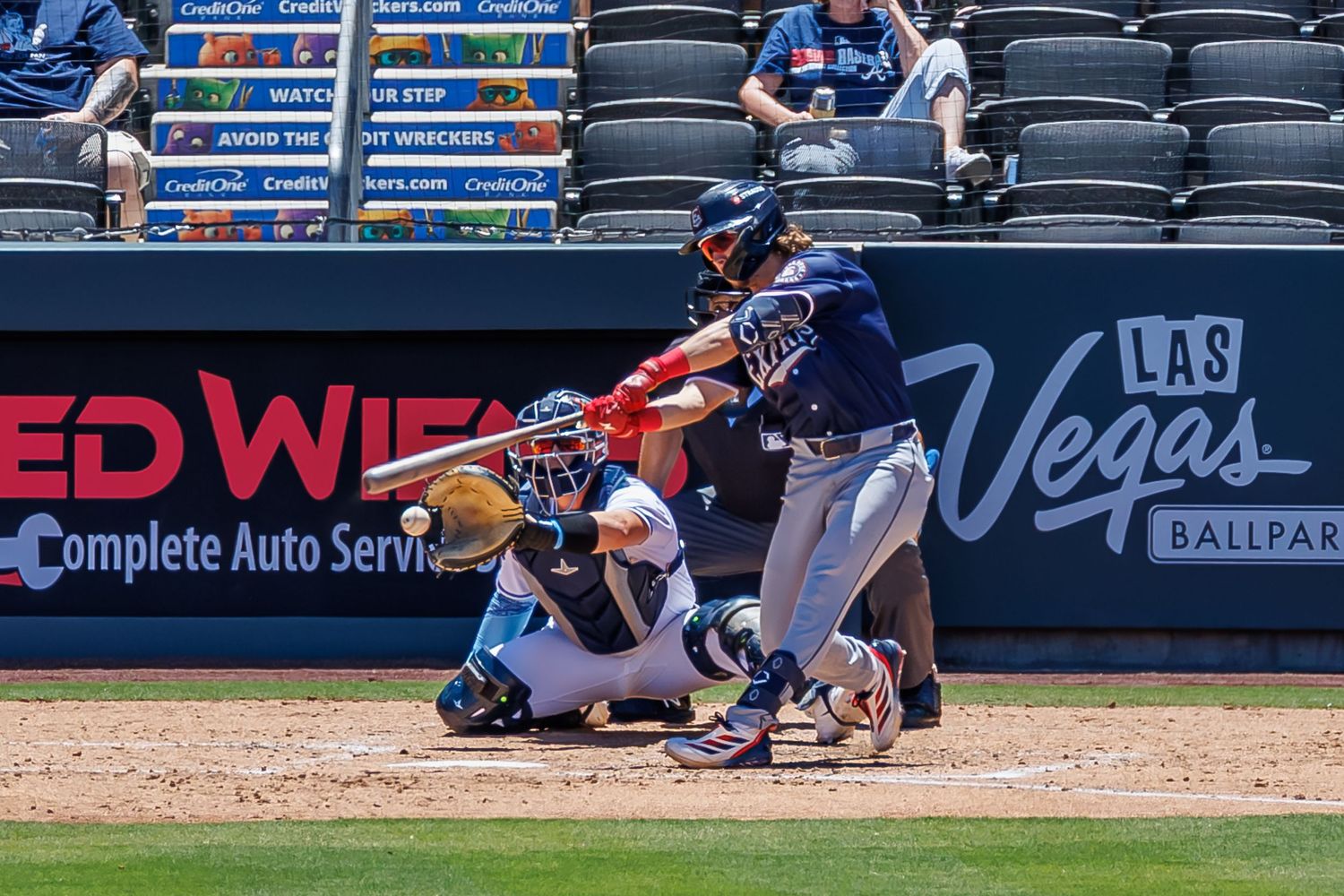Baseball batter swinging at pitch with catcher crouching behind home plate at Las Vegas Ballpark on sunny day.