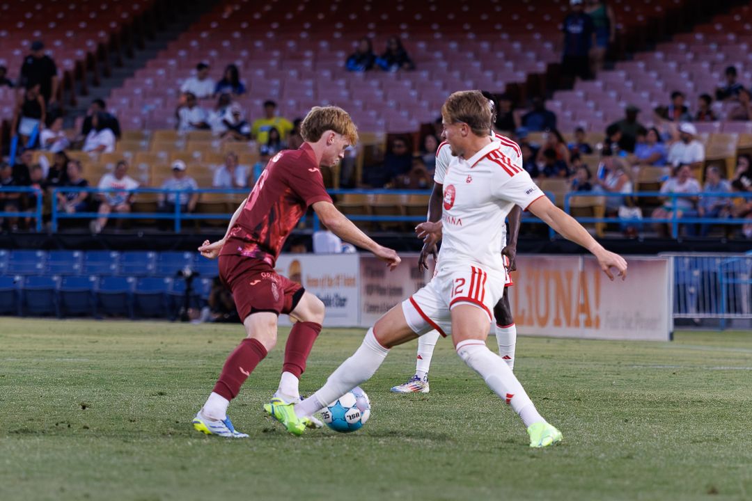 Two soccer players compete for the ball during a night match, one in dark red and one in white uniform.