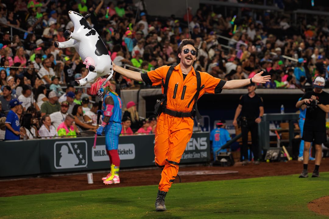 Man in orange jumpsuit with arms outstretched runs onto baseball field during a lively stadium event at night.