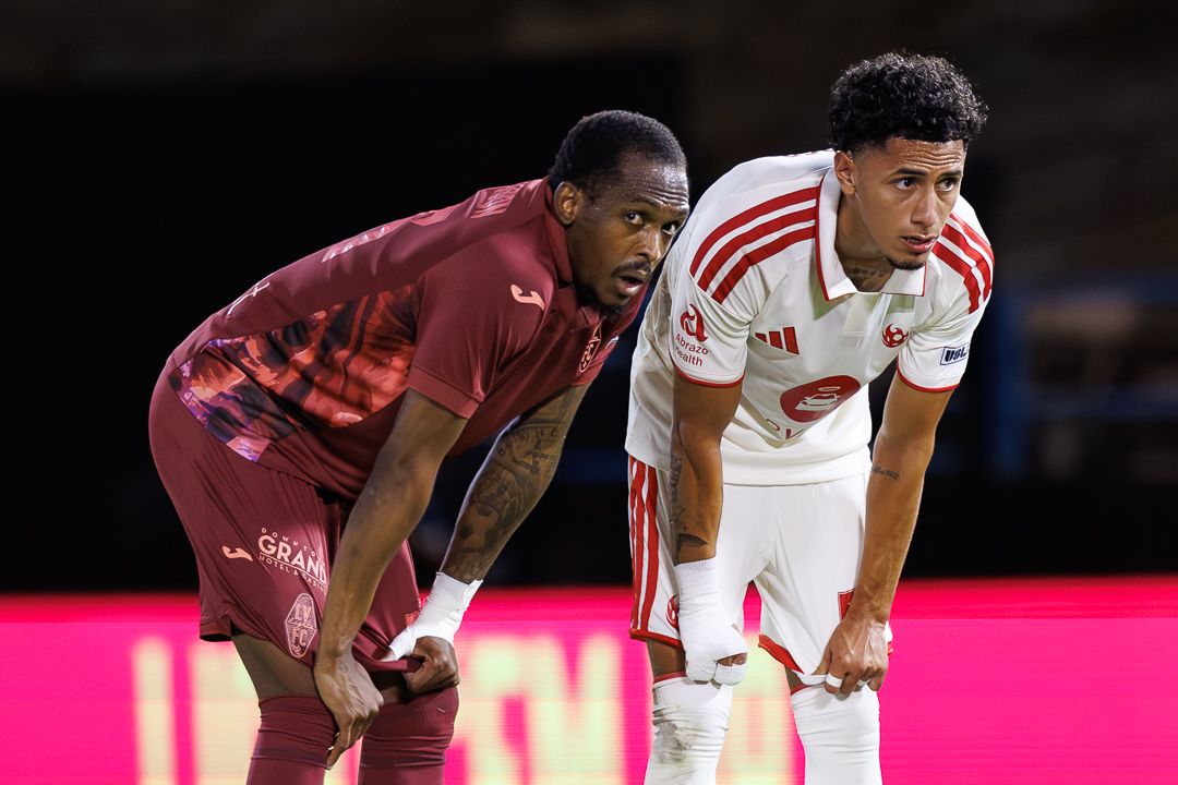 Two soccer players in red and white uniforms crouch on the field during a competitive match.