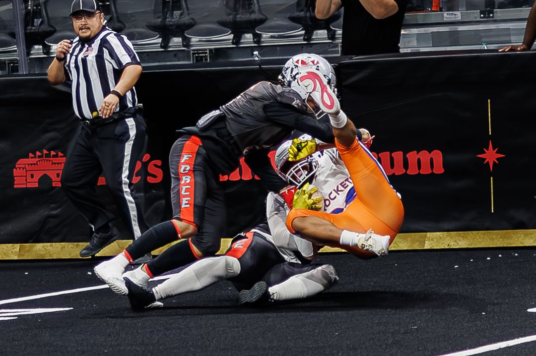 Two arena football players collide during an intense indoor game, with a referee observing the action nearby.