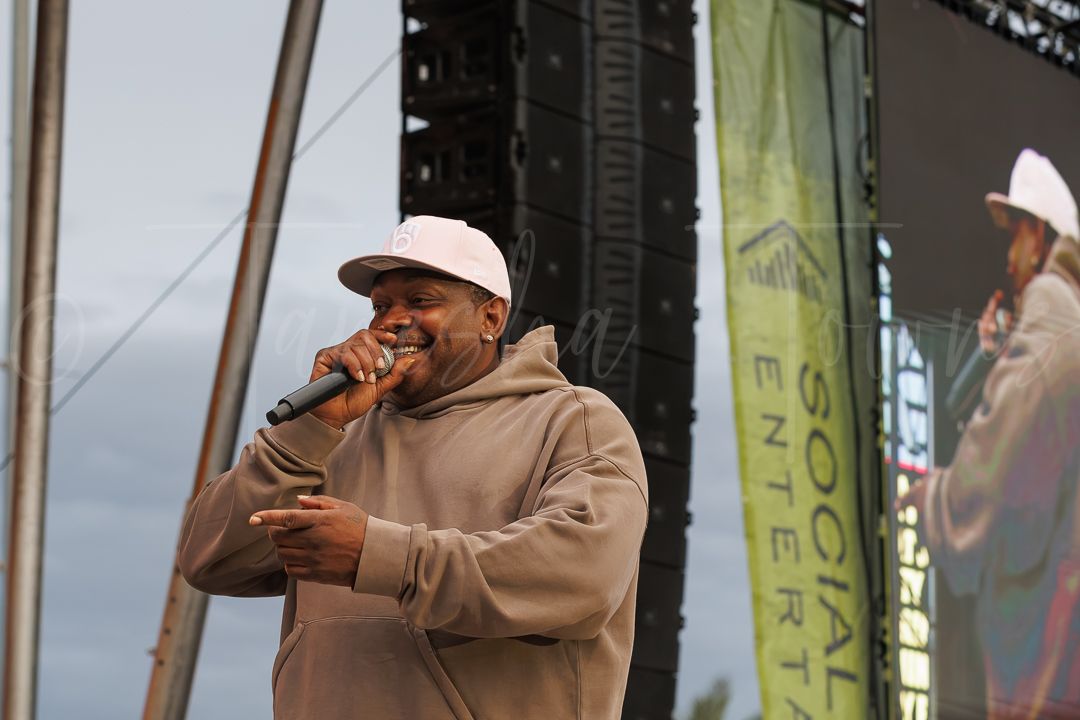 Man in beige hoodie and cap performing on outdoor stage with microphone at a live music festival event.