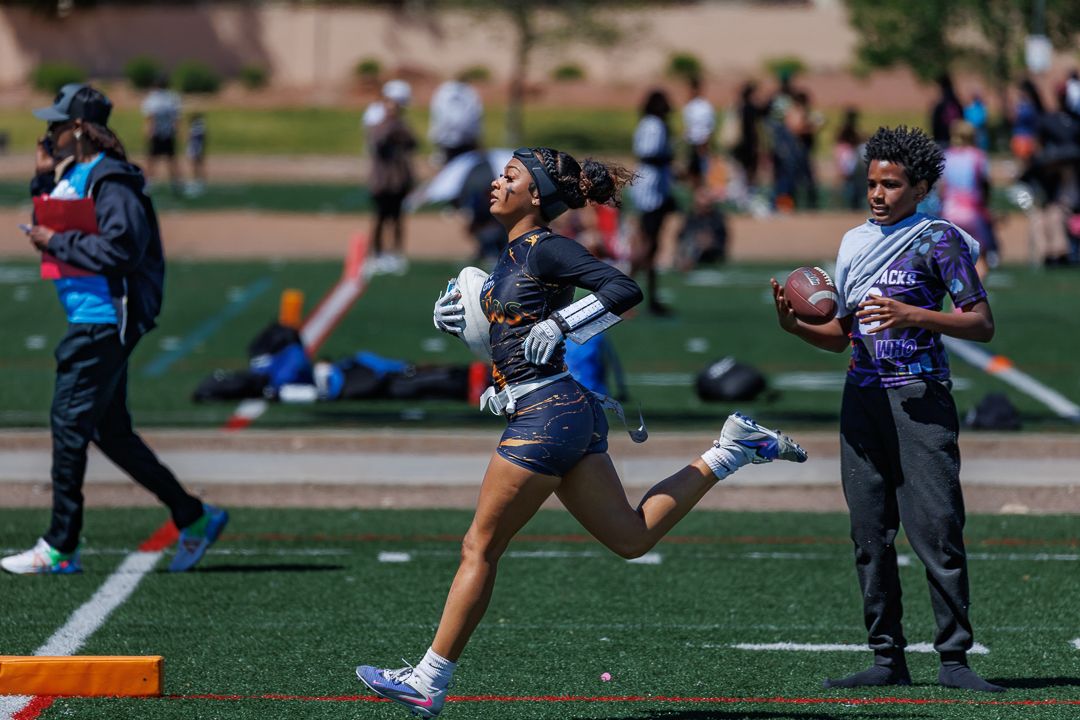 Female athlete sprints with football during outdoor flag football game on green turf field with players in background.