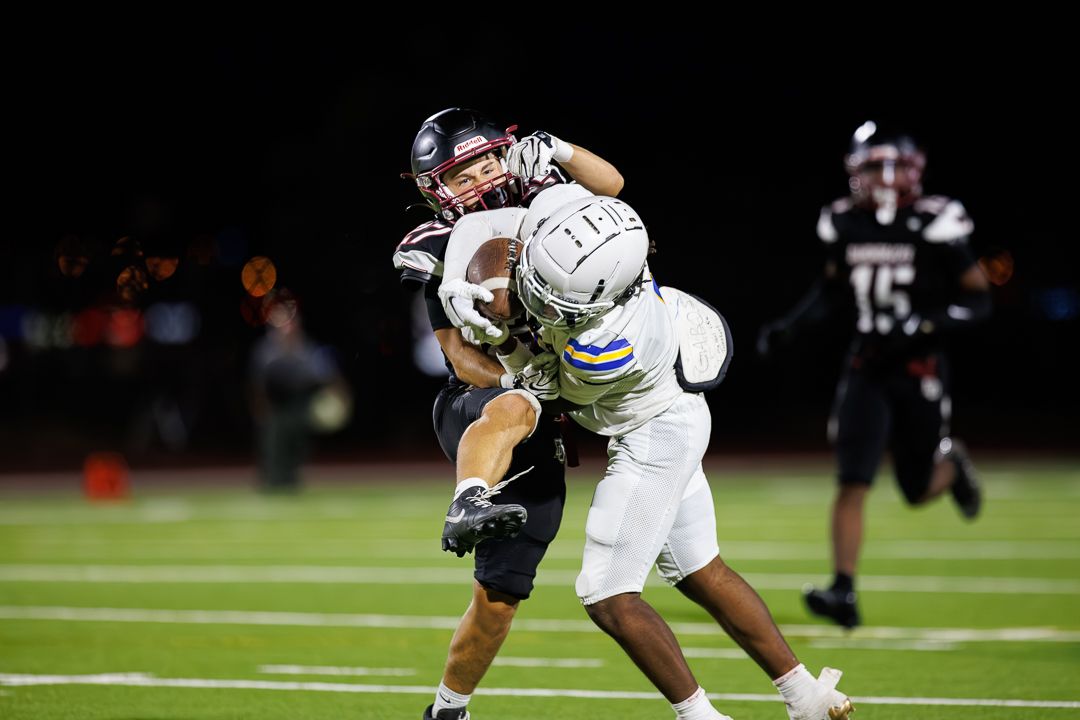 Two football players collide as one attempts to catch the ball during a night game under stadium lights.