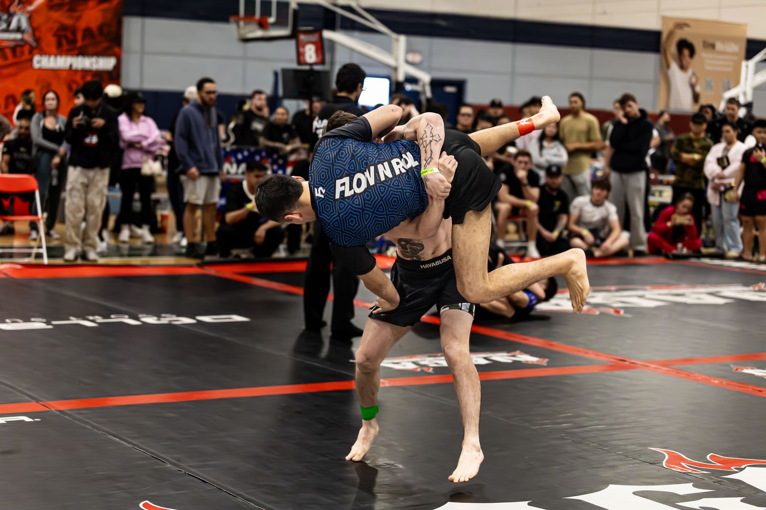 Two wrestlers compete at a grappling championship, one executing a powerful takedown throw on a red and black mat.