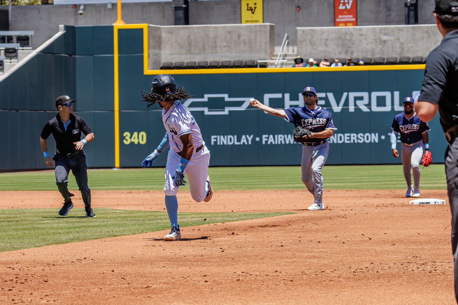 Baseball players in action on the field, with a runner rounding a base during a sunny daytime game.