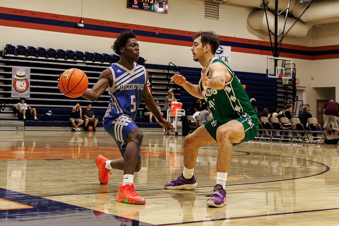 A basketball player in blue dribbles past a defender in green during an indoor college basketball game.
