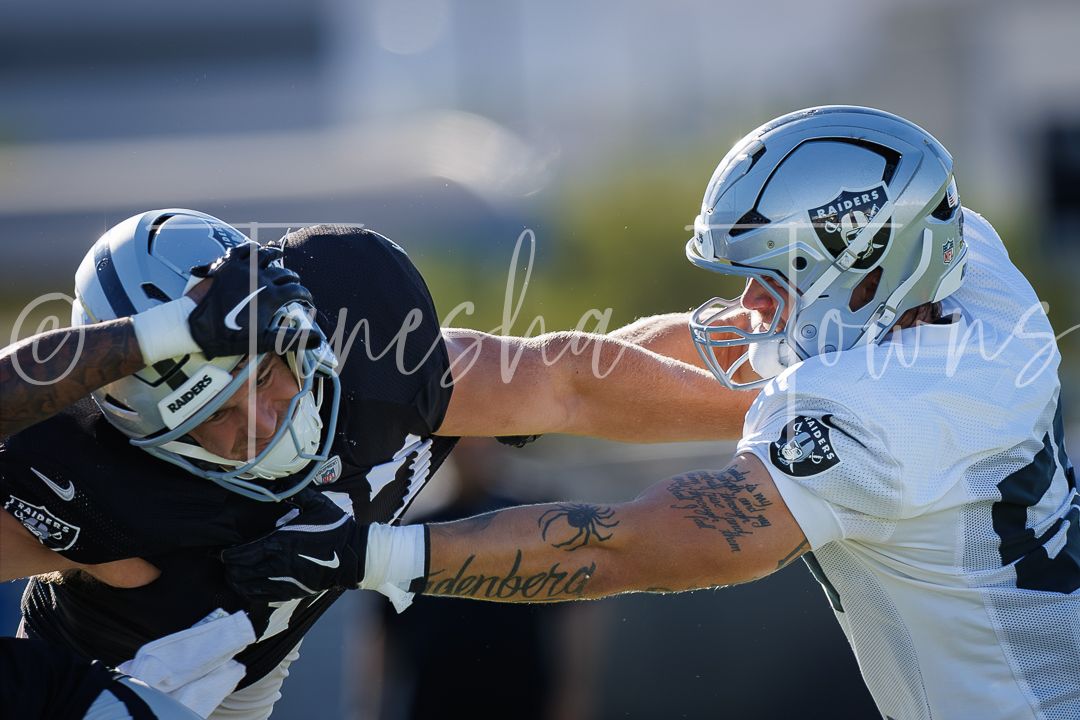 Two football players engage in a blocking drill during practice, one in dark and one in white uniform.