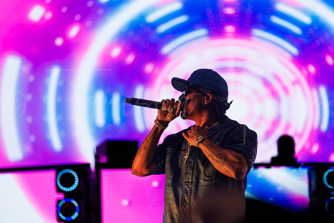 Male rapper performing on stage with microphone, wearing cap, in front of vibrant pink and blue tunnel light display.