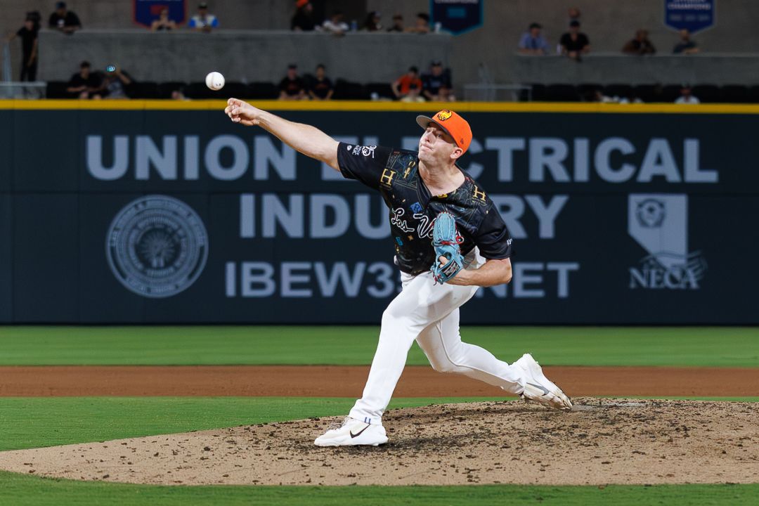 Minor league baseball pitcher in navy uniform throws a pitch at night with Union Electrical Industry signage behind.