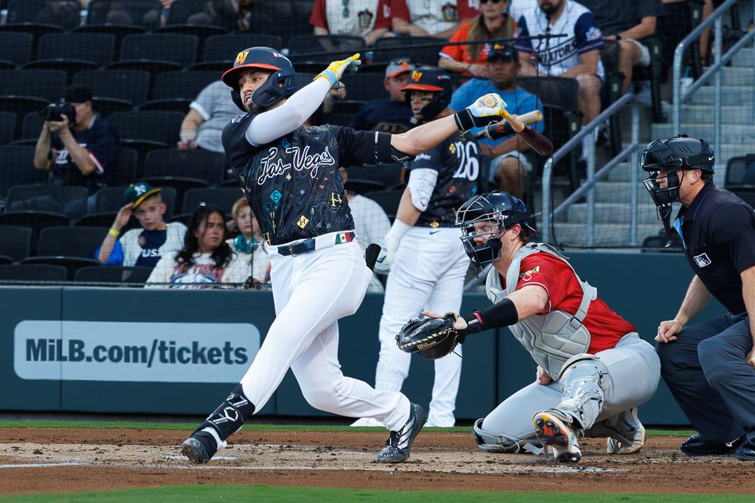 Minor league baseball batter swings at pitch with catcher and umpire behind home plate during MiLB game.