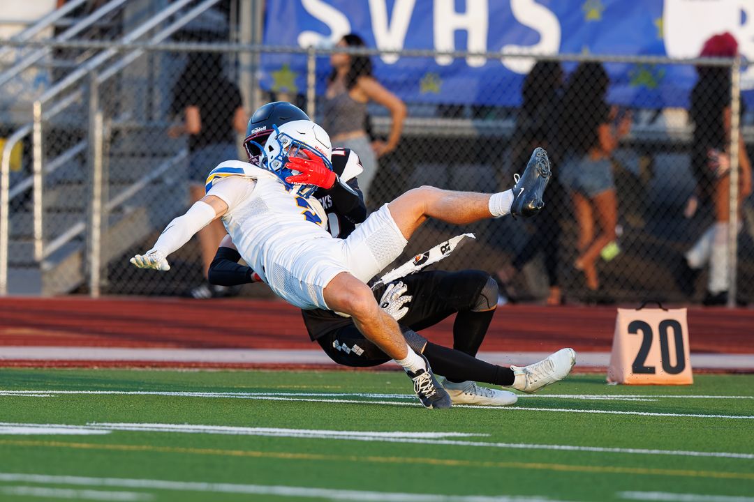 High school football player in white uniform dives to make a catch near the 20-yard line during a game.