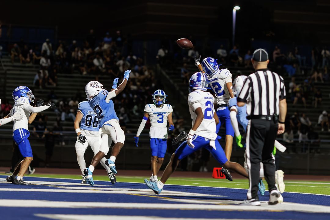 High school football players compete in a night game, with a player leaping to catch a pass in the end zone.