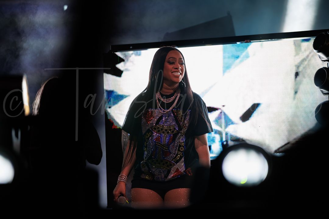 Female performer on stage viewed through gap in curtains wearing graphic tee and shorts with layered necklaces.