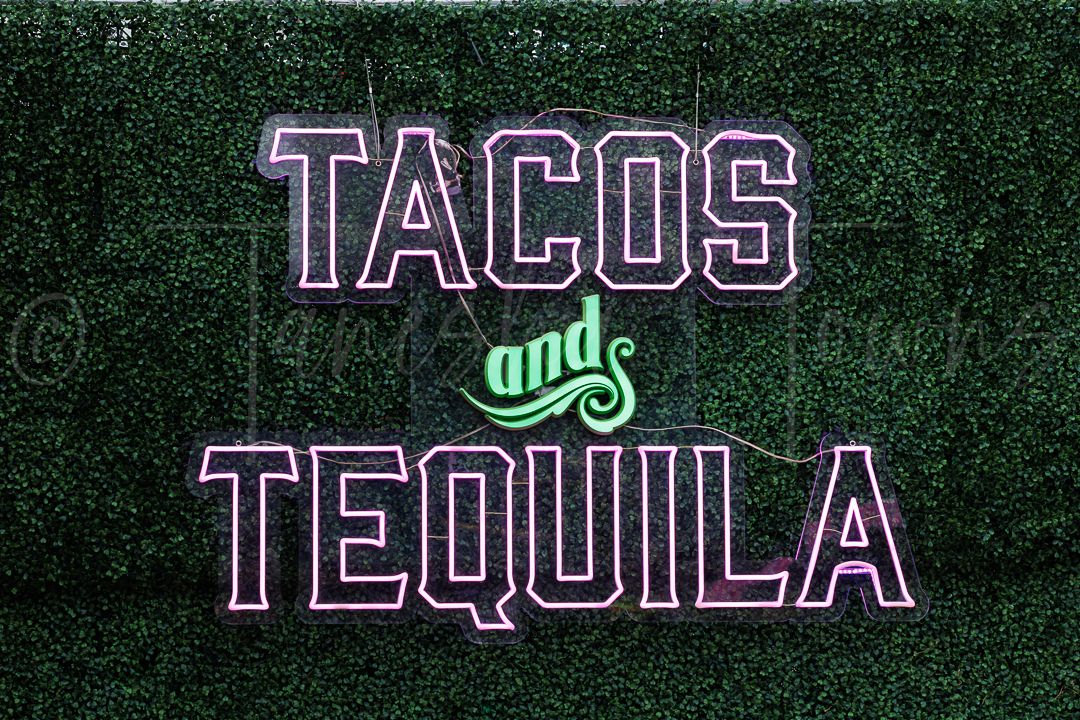 Tacos and Tequila neon sign on green hedge wall backdrop.