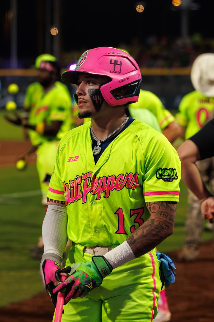 Player #13 in neon green Chili Peppers jersey and pink helmet #44 holds bat during night softball game.