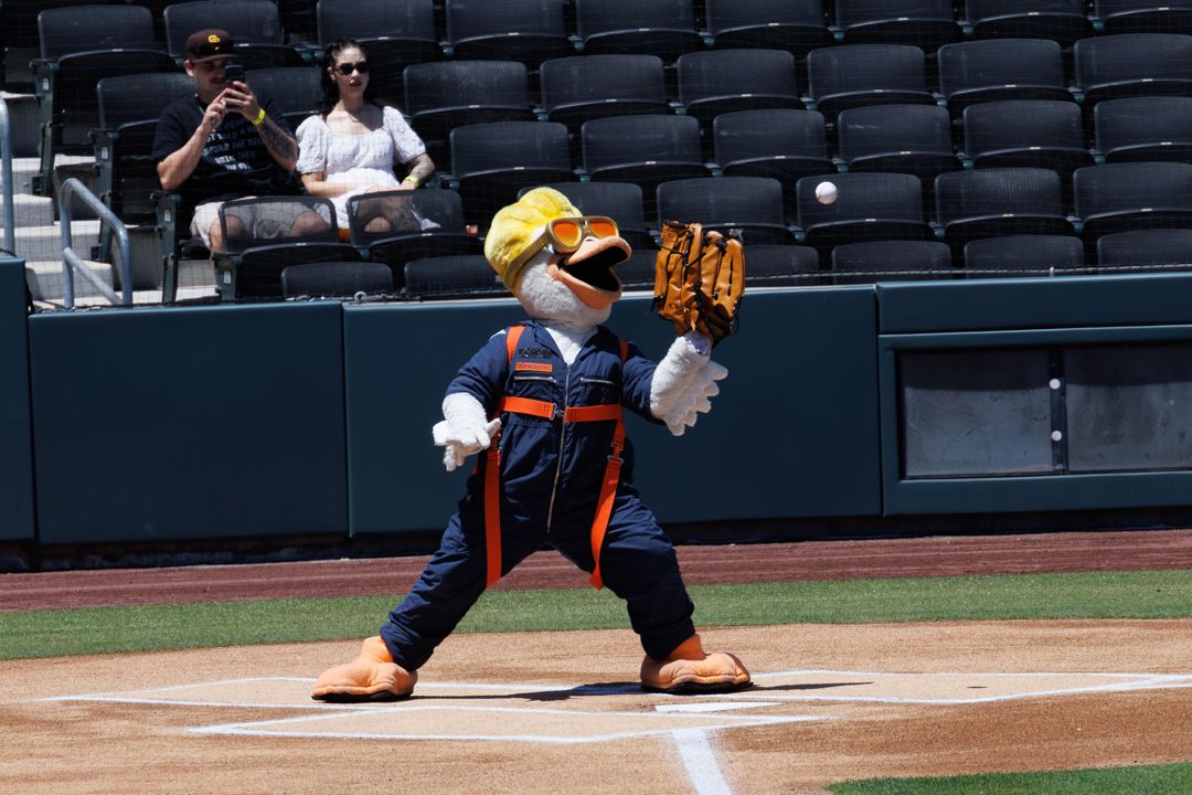 A baseball mascot in orange uniform and yellow helmet catches with a glove at home plate during a sunny game day.