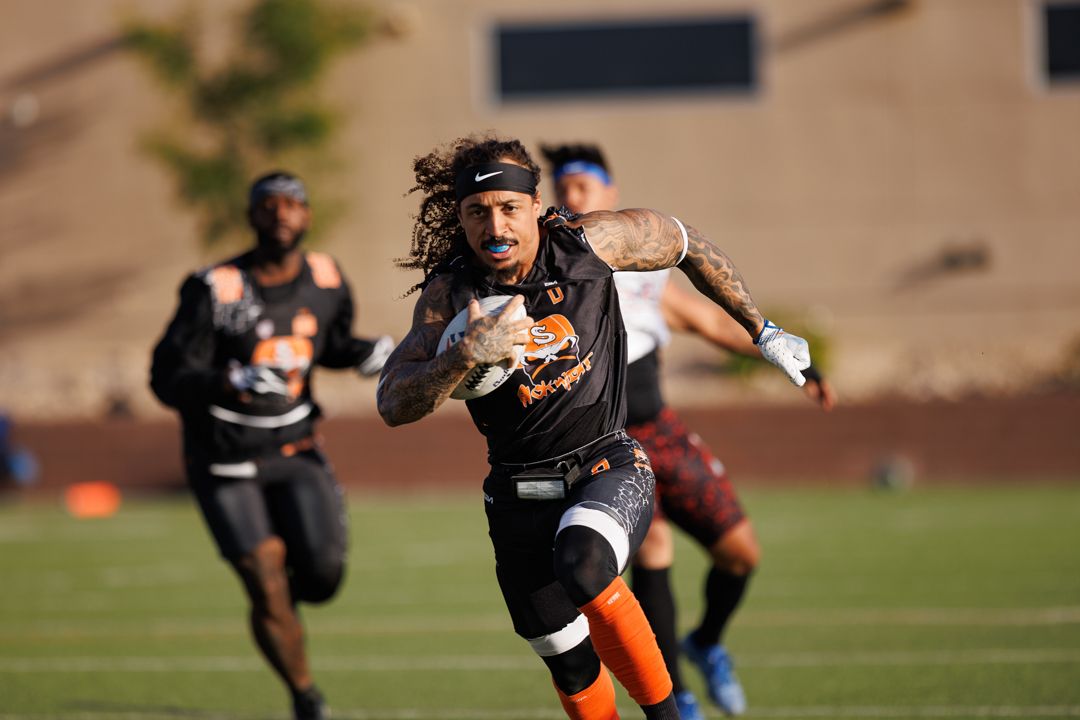 Football player with long hair runs with the ball during an outdoor flag football game in orange and black uniform.