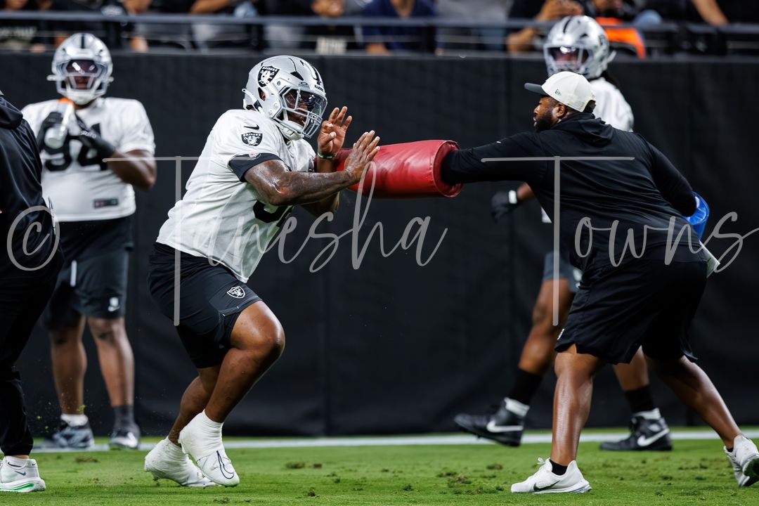 NFL player in black Raiders uniform practices blocking drills with a coach holding a red pad during training camp.