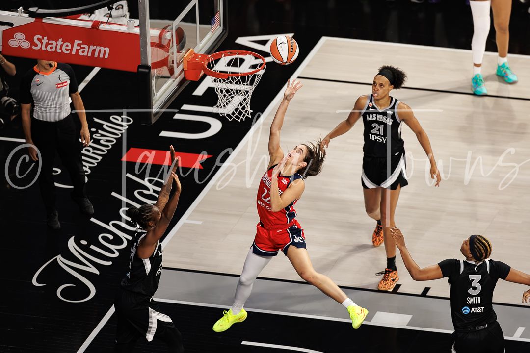 WNBA player drives to the basket during a game while defenders attempt to block the shot under the hoop.