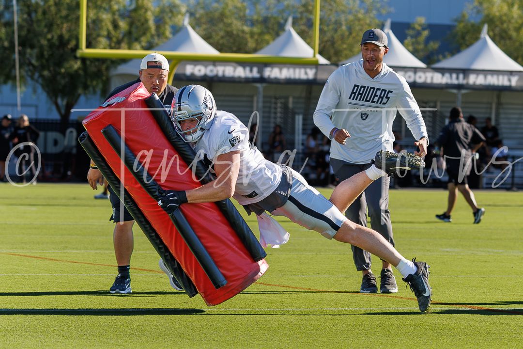 Raiders football player drives through a blocking pad during team practice drill on a sunny day.