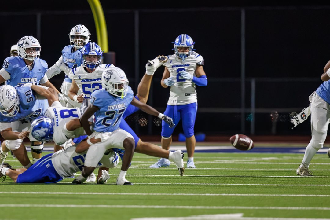 Football players in blue and white uniforms compete during a night game, with a loose ball visible mid-play.