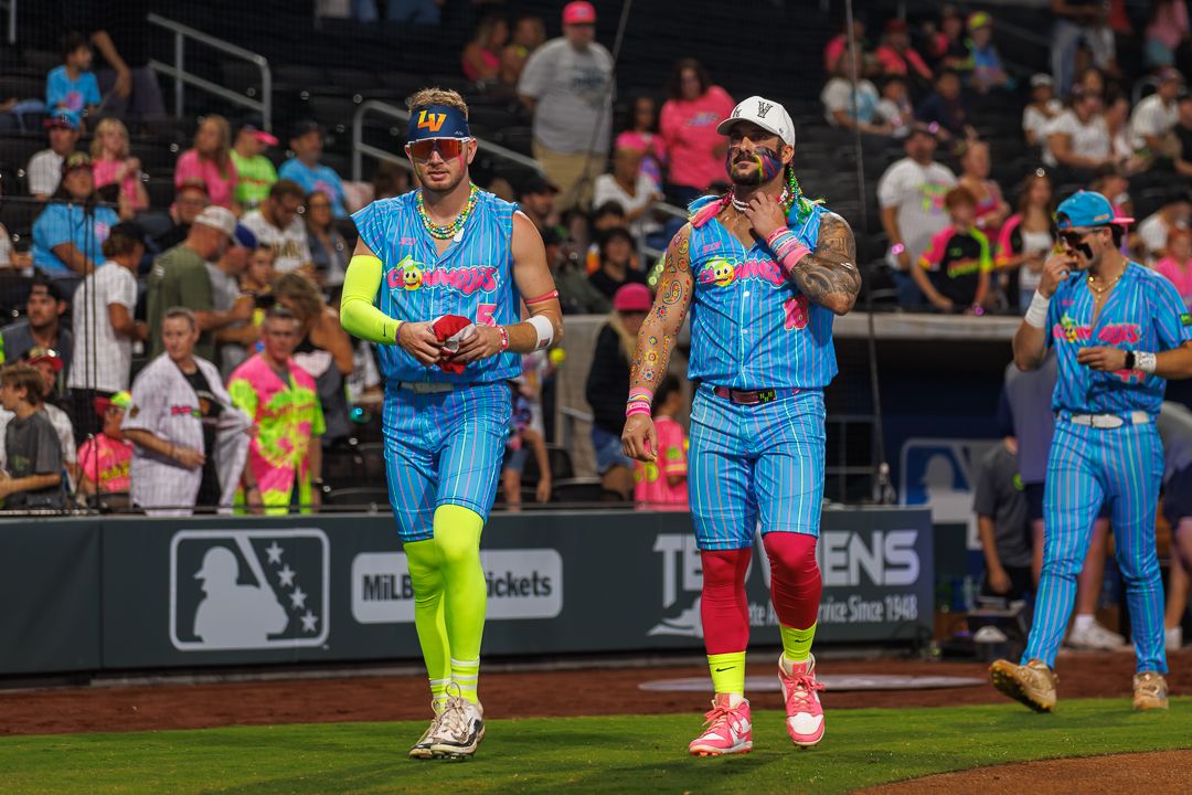 Two baseball players in colorful neon uniforms walk on field during MLB celebrity softball game.