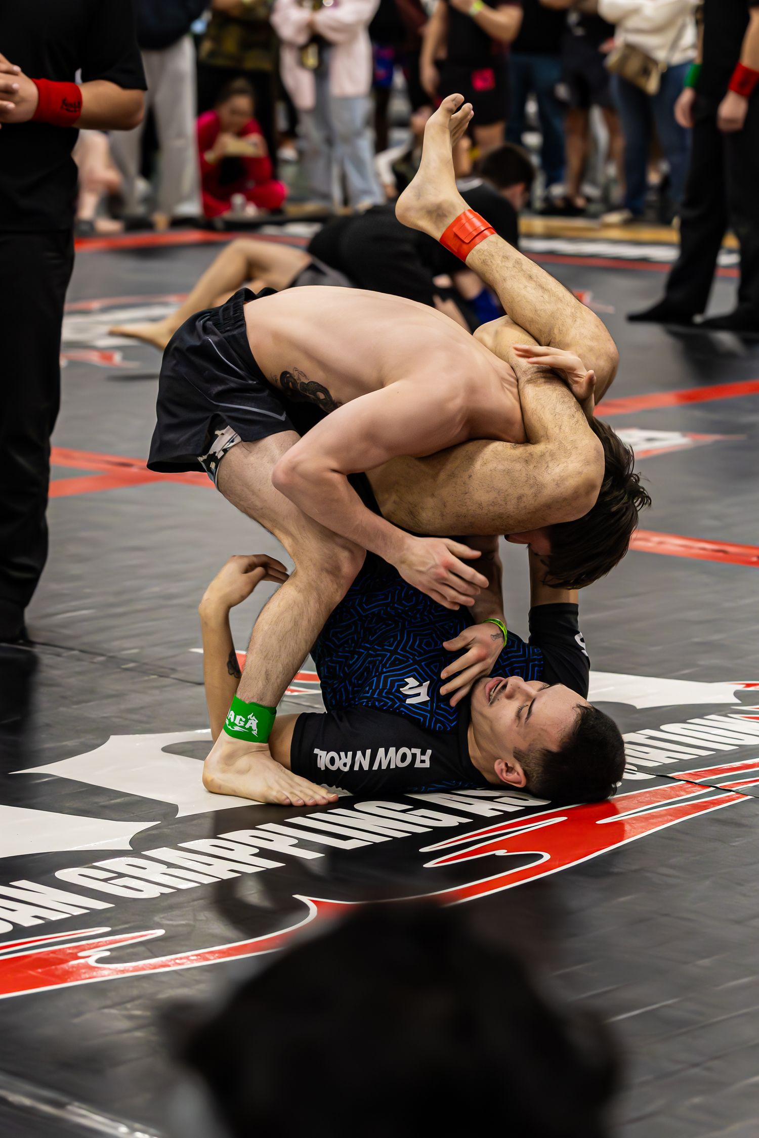 Two competitors grappling on a mat at a Flow N Roll BJJ tournament during an intense ground fighting match.