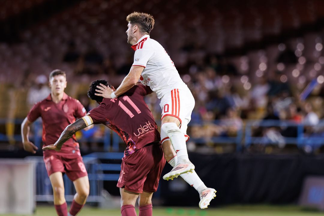Soccer players compete for a header during an evening match, with a packed stadium in the background.