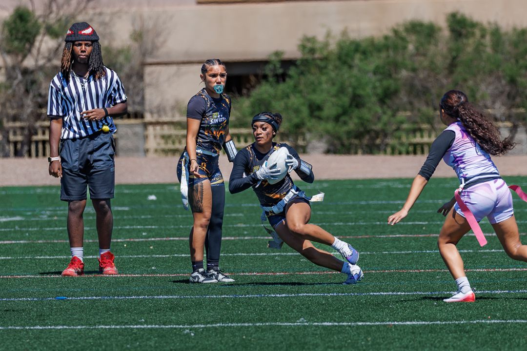 Female flag football players compete on a green turf field with a referee observing the play during a sunny outdoor game.