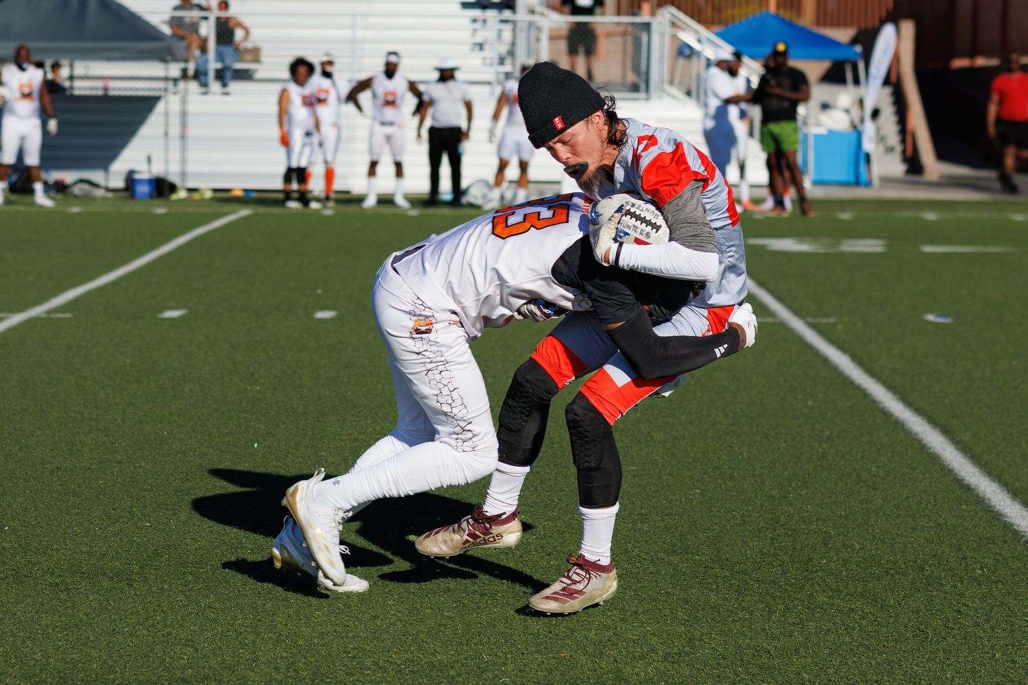 Two football players in orange and white uniforms practice blocking drills on a blue turf field during a sunny day.