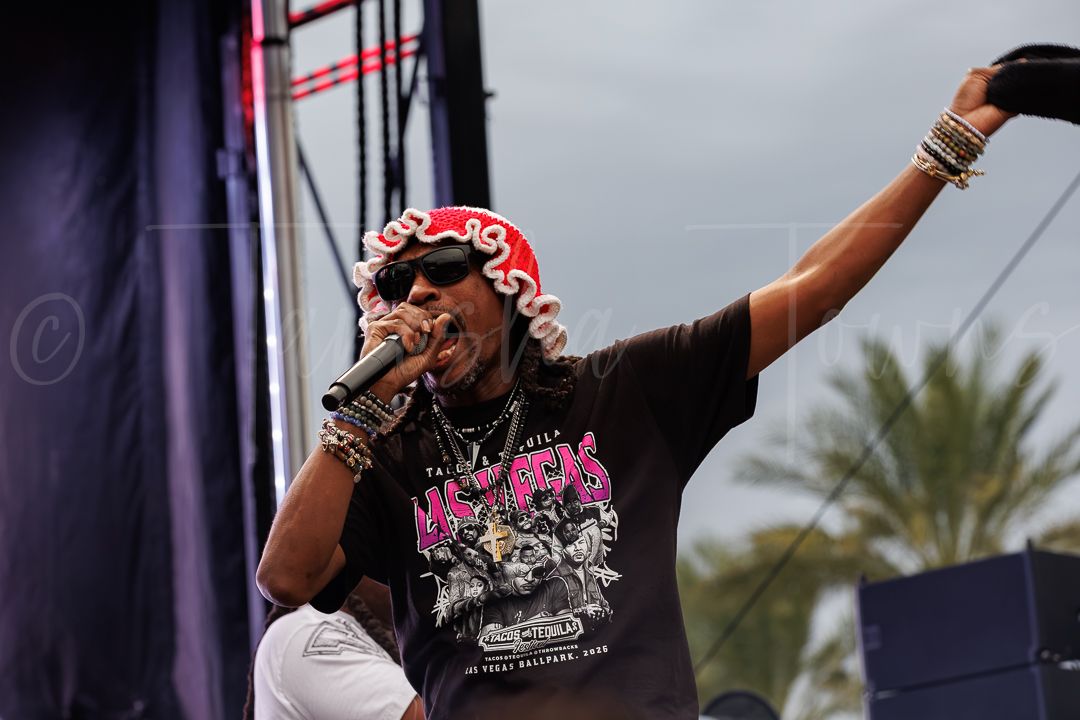 A male rapper performs on stage outdoors, wearing a graphic tee and red cap, raising his fist while holding a microphone.