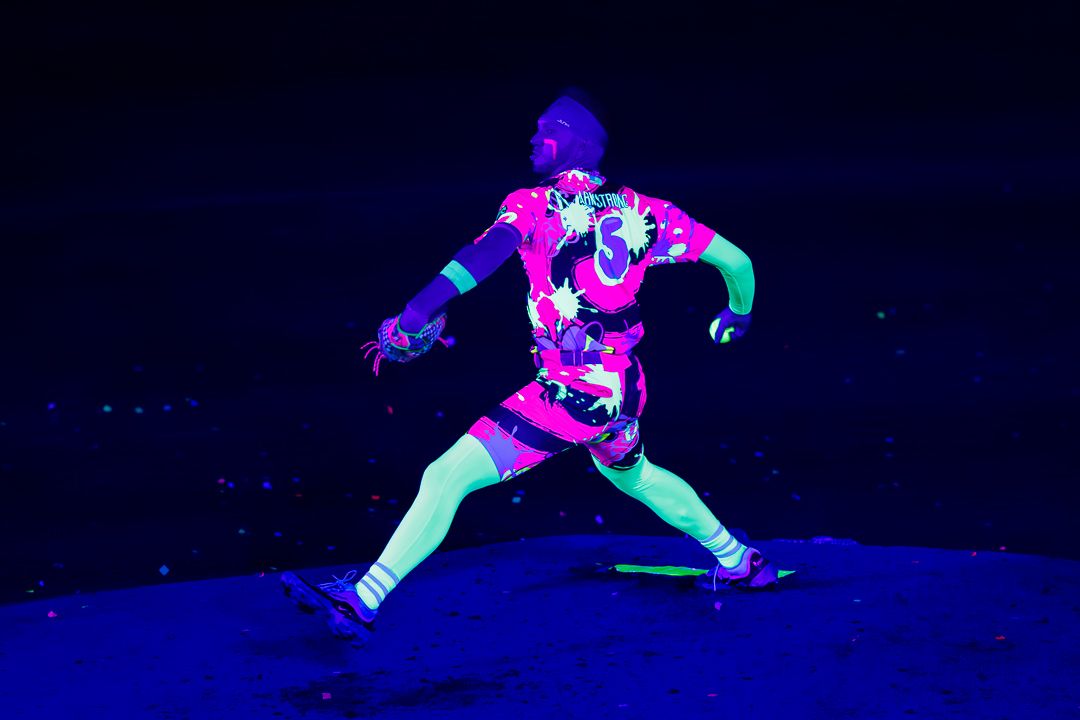 Person in glowing neon pink floral outfit pitching under UV blacklight during a vibrant nighttime sports event.