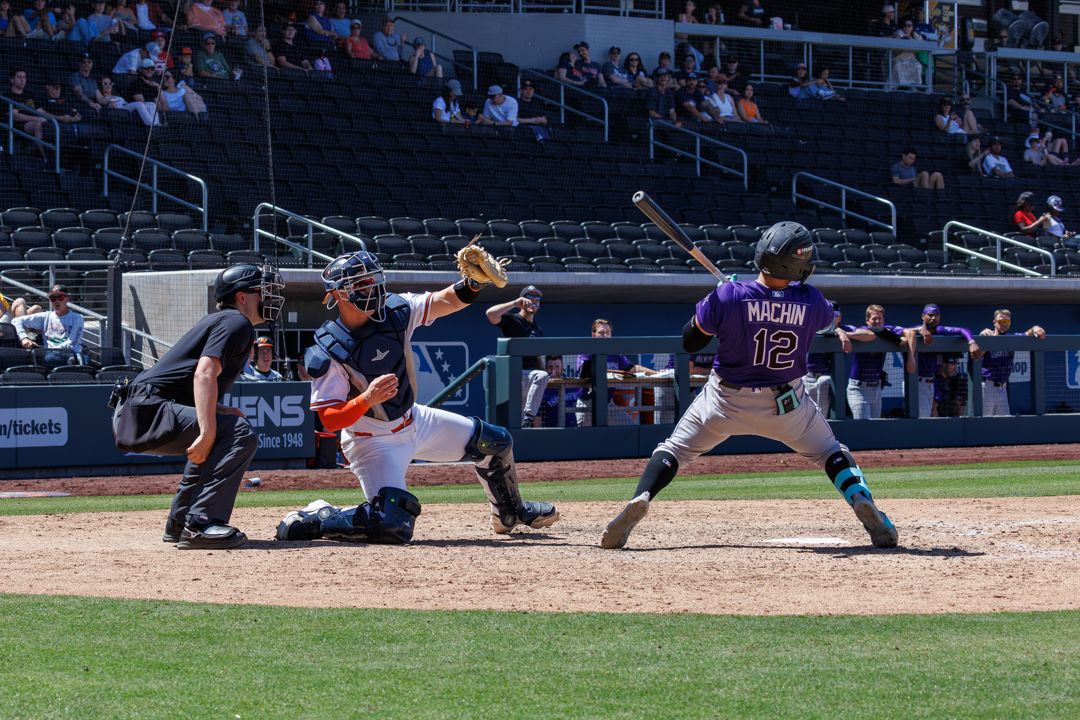 Baseball batter in purple uniform prepares to swing during daytime game with catcher and umpire.