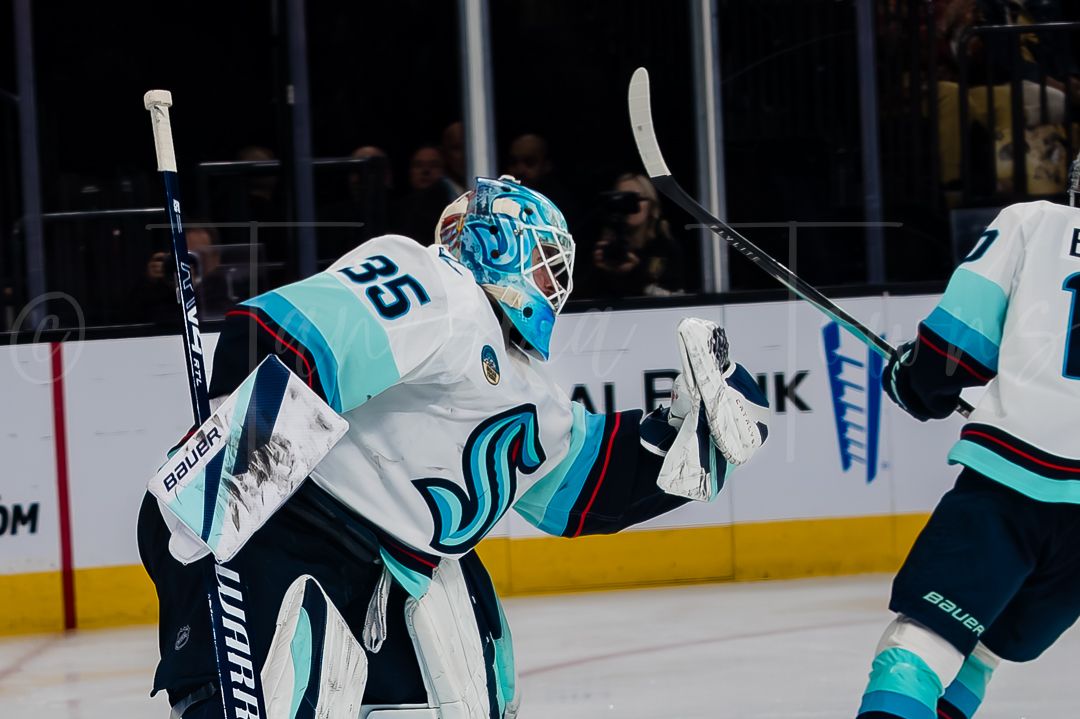 A hockey goalie in teal and white uniform stands ready in the crease during a game.