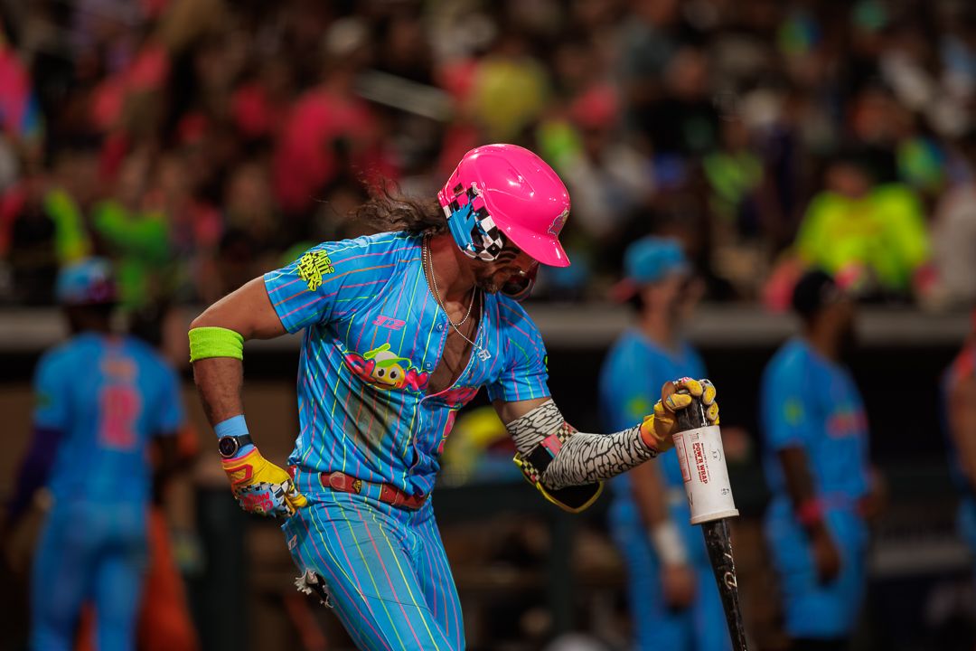 Cricket batter in bright pink helmet and blue uniform prepares to swing during a colorful night match.