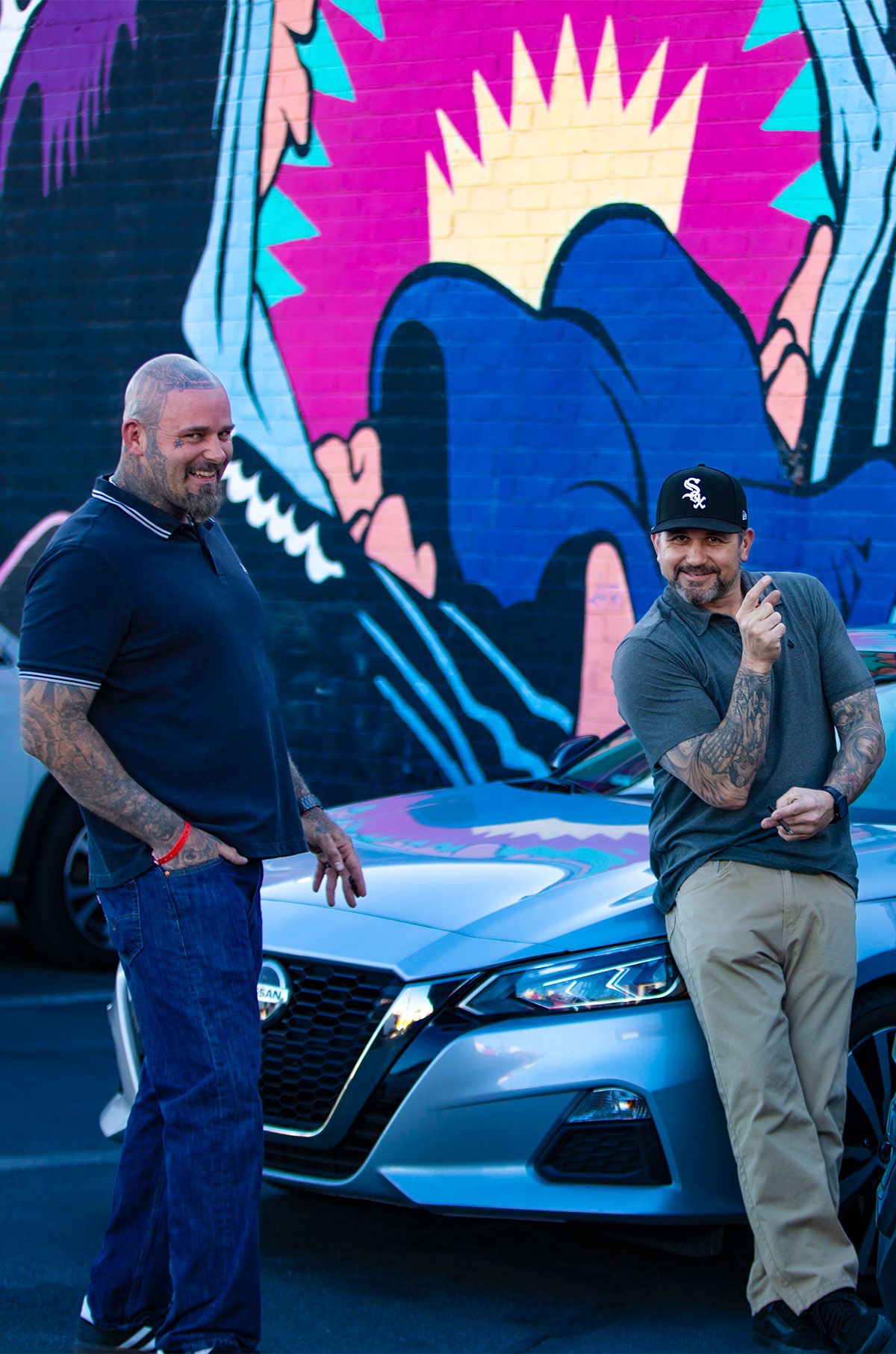 A silver car parked in front of a vibrant pink and blue graffiti wall.