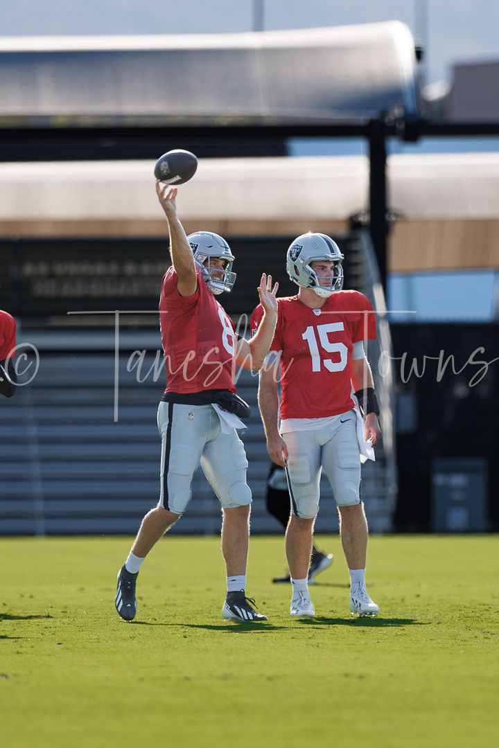 Two Las Vegas Raiders quarterbacks in red jerseys practice throwing drills on an outdoor field.