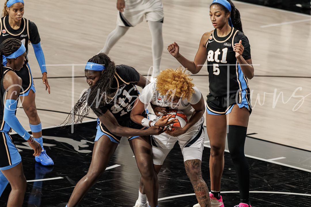 WNBA players battle for possession during an intense basketball game, with players in light blue and white uniforms competing.