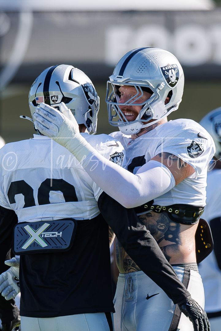 Two Las Vegas Raiders NFL players in white uniforms and silver helmets embrace on the field during a game.