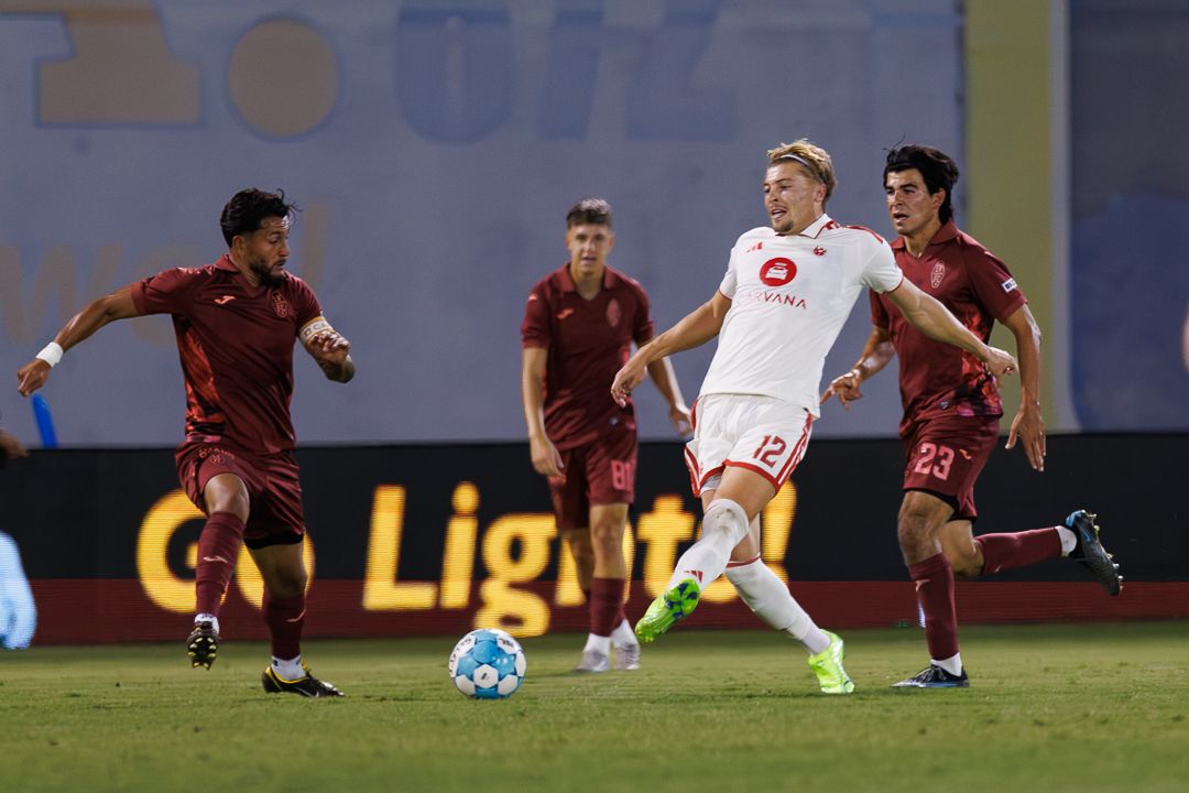 A blonde soccer player in white dribbles past defenders in red during a night match on a professional field.