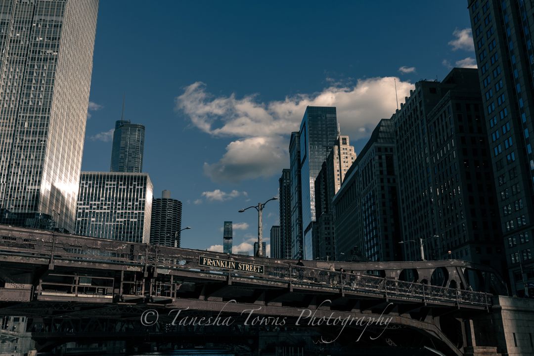 Panoramic cityscape view of modern skyscrapers and buildings along a waterfront with blue sky and white clouds overhead.